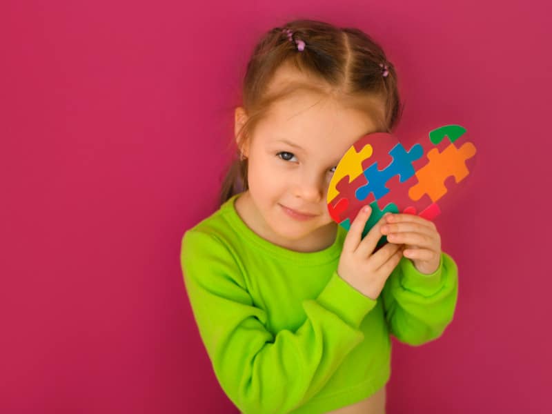 menina com uma blusa verde sorrindo e segurando um coração com as cores do autismo