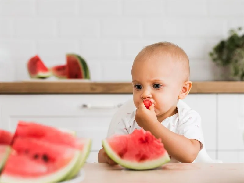 bebê sentado comendo uma fatia de melancia com a mão 