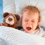 Criança chorando na cama com um ursinho de pelúcia marrom ao lado