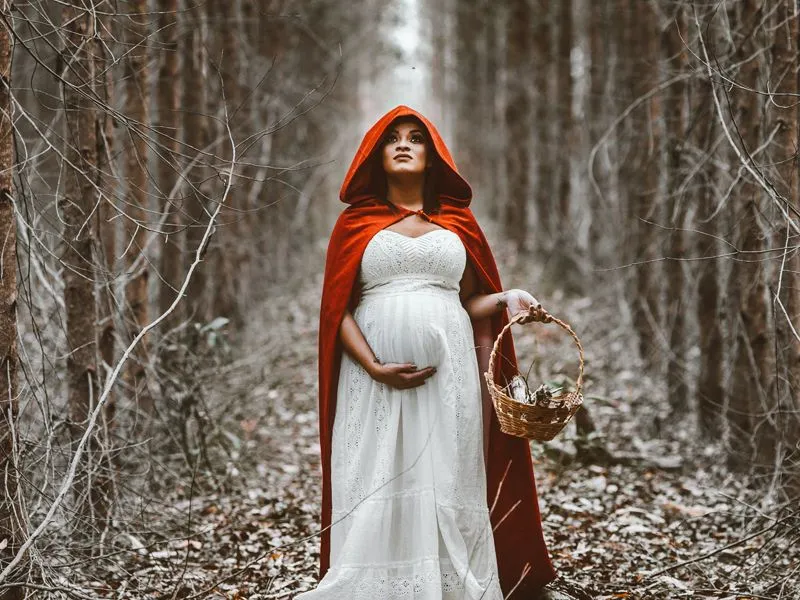 Mulher grávida em floresta com roupa de chapeuzinho vermelho.