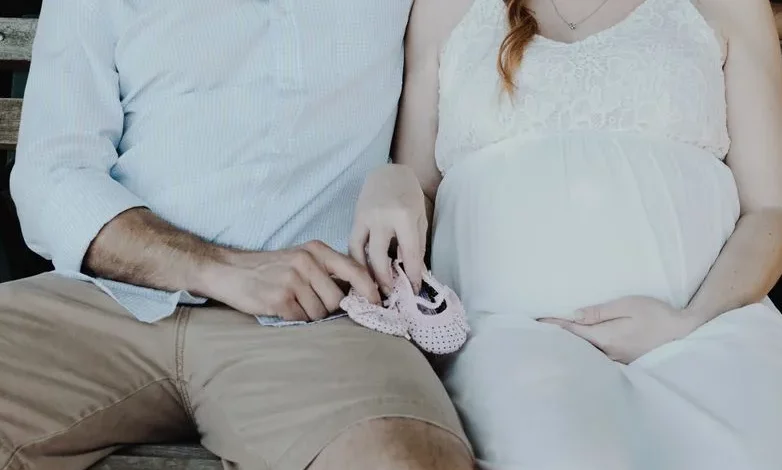 Homem e mulher grávida sentados segurando sapatinho rosa de crochê.