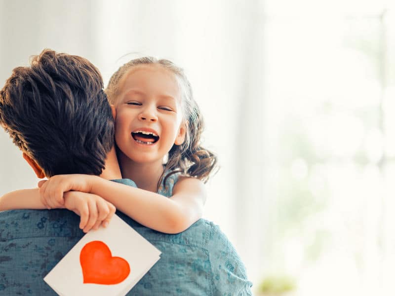 pai abraçando a filha sorrindo com um cartão de Dia dos Pais na mão