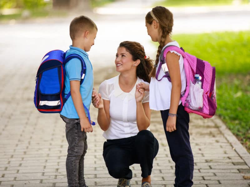 uma mãe, agaixada e conversando com os seus dois filhos um menino de bolsa azul e uma menina de bolsa rosa