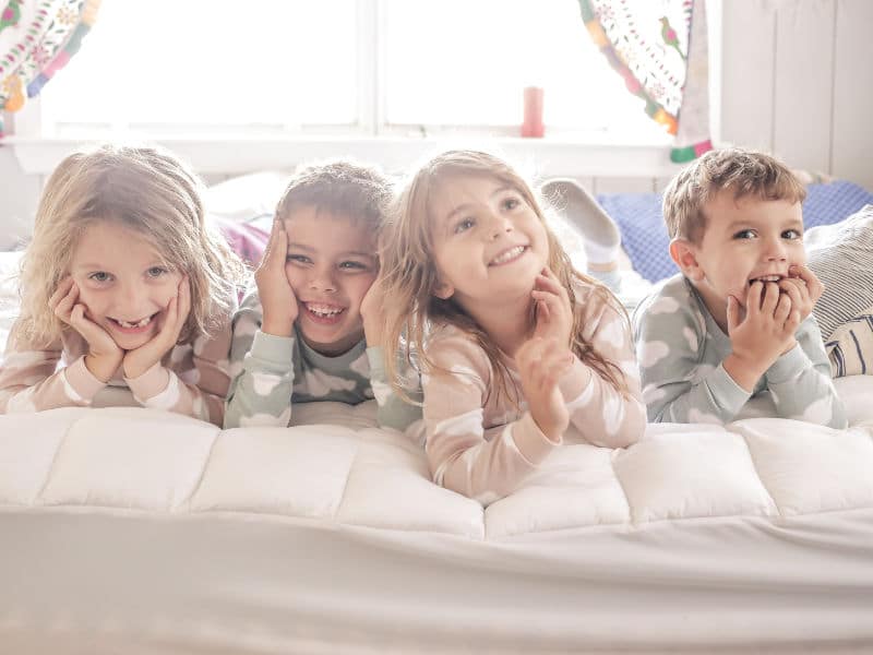 duas meninas e dois meninos brancos deitados na cama felizes em uma festa do pijama