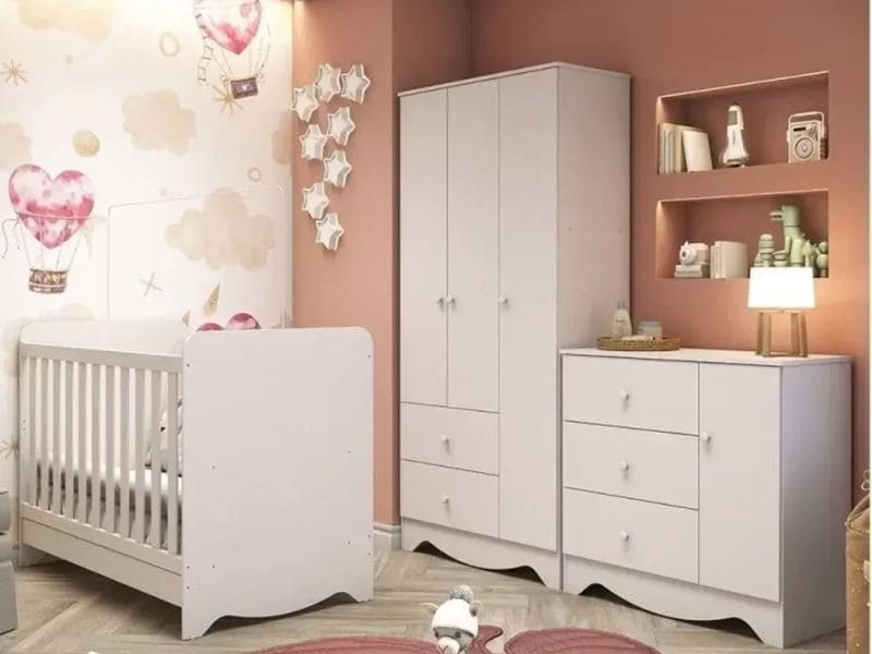  Quarto de bebê com berço branco, guarda-roupa, cômoda e prateleiras de parede. A decoração de quarto de bebê inclui paredes adornadas com balões de ar quente e nuvens. Um brinquedo macio repousa sobre o tapete com padrão chevron.