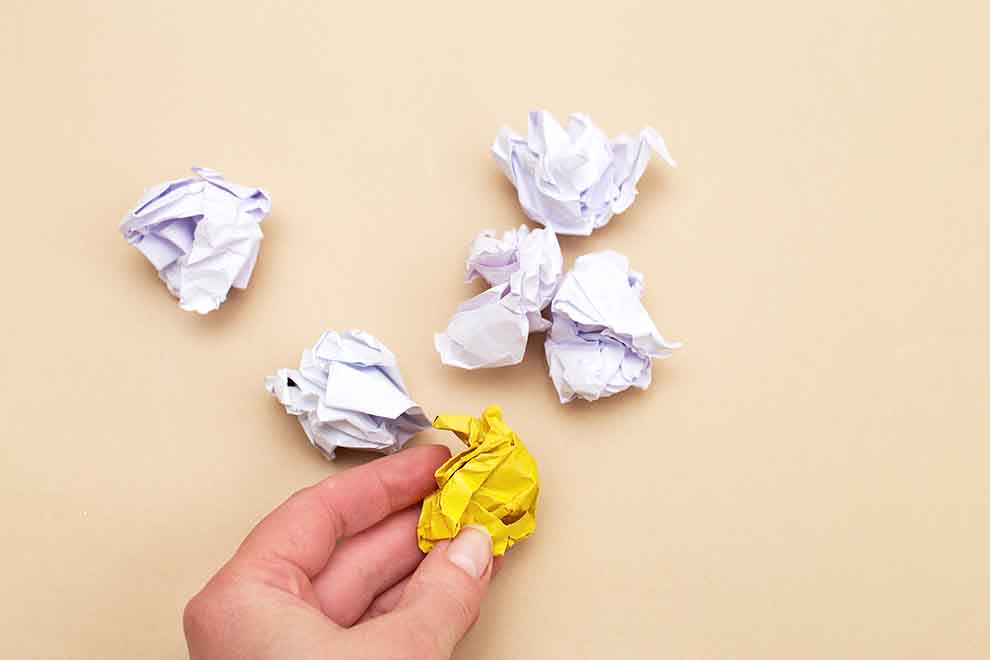 Mão segurando papel amassado amarelo entre papéis brancos.